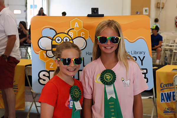 Two smiling girls wearing sunglasses and green award ribbons stand in front of a colorful Harford County Public Library Spelling Bee sign at the Harford County Farm Fair. One girl wears a red shirt and the other a pink shirt with a unicorn patch.
